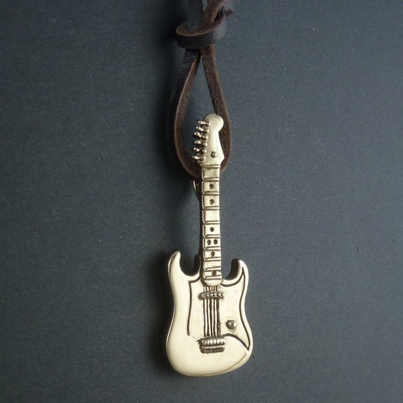 May include: A gold-colored electric guitar pendant on a brown leather cord. The guitar has a white body and a detailed fretboard.