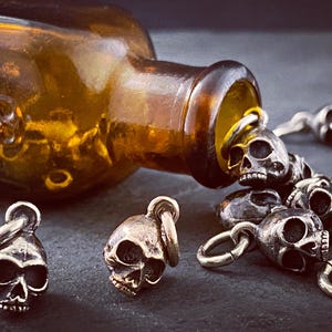 May include: A collection of silver skull charms spilling out of an amber glass bottle with a skull and crossbones design.