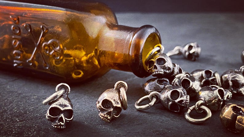 May include: A collection of silver skull charms spilling out of an amber glass bottle with a skull and crossbones design.