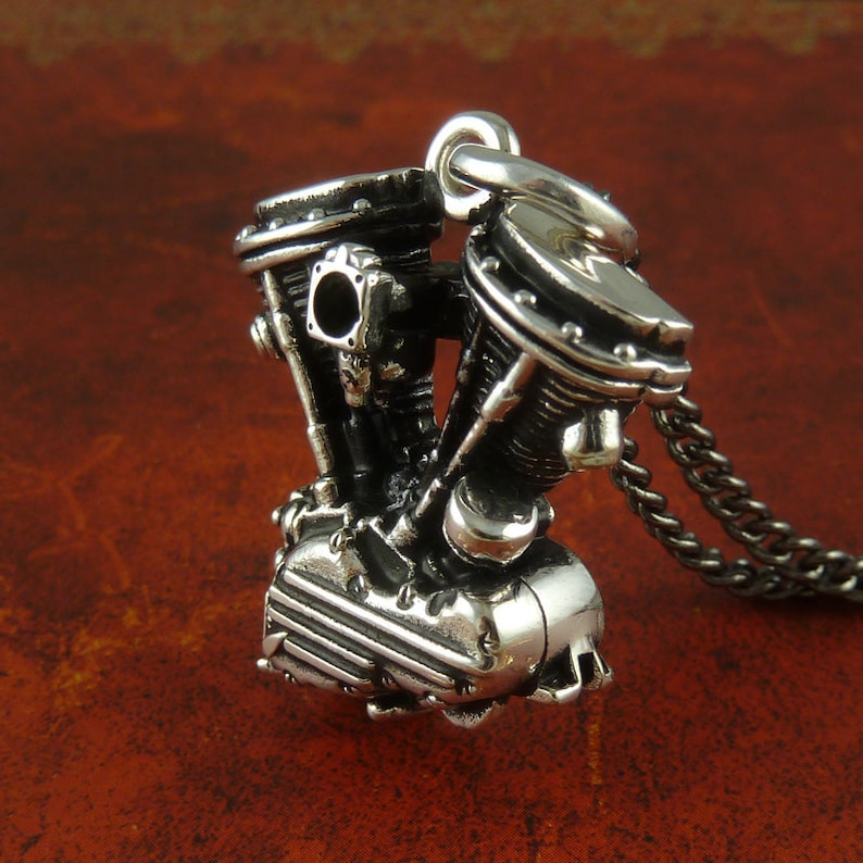 May include: A silver pendant shaped like a motorcycle engine. The pendant is intricately detailed with carvings and has a loop at the top for attaching a chain.