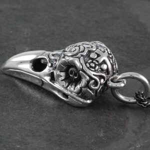 May include: Silver pendant featuring a detailed, ornate raven skull with a loop for attaching a chain. The skull is intricately designed with floral and scrollwork patterns.
