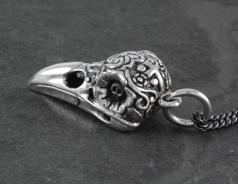 May include: Silver pendant featuring a detailed, ornate raven skull with a loop for attaching a chain. The skull is intricately designed with floral and scrollwork patterns.