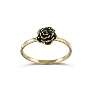 Puede incluir: Un anillo dorado con un diseño de rosa oscura. El anillo tiene una banda delgada y una flor de rosa detallada en el centro. Es una pieza de joyería.