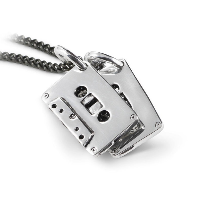 May include: A silver-colored cassette tape pendant with a black chain. The pendant is rectangular with detailed features, including holes and a central tape window. The chain is a dark, textured metal, and the background is white.