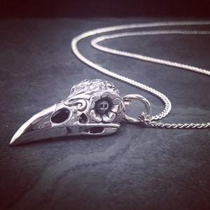 May include: A silver pendant necklace featuring a detailed, ornate skull with a floral design. The skull has a small skull detail in the eye socket. The pendant is attached to a silver chain.