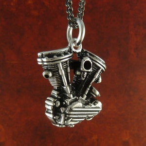 May include: A silver pendant shaped like a motorcycle engine. The pendant is intricately detailed with carvings and has a loop at the top for attaching a chain.