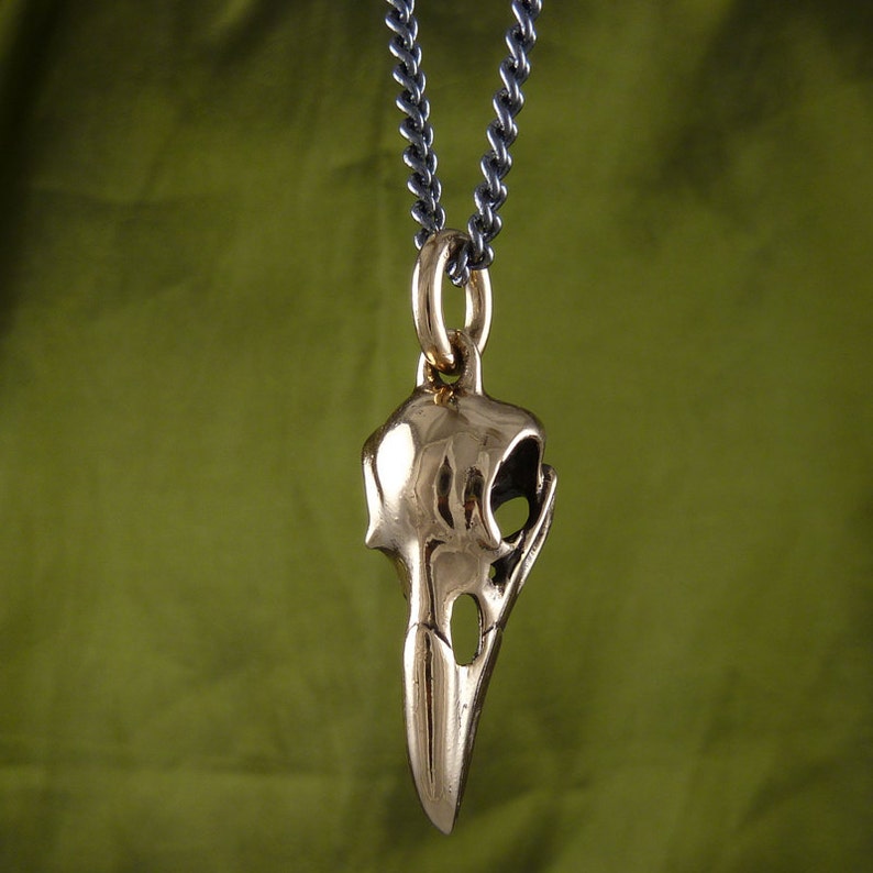 May include: A gold-colored bird skull pendant on a silver chain. The pendant is detailed and realistic, with a textured surface and visible eye sockets.
