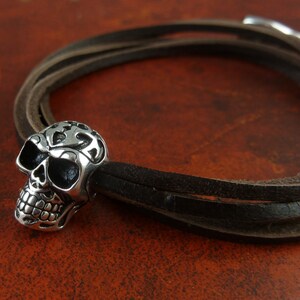 May include: A brown leather bracelet with a silver skull charm. The skull has intricate details and a black eye socket.