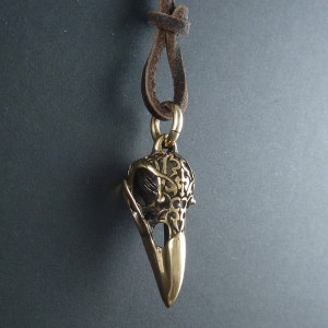 May include: A gold-toned pendant necklace featuring a detailed, ornate bird skull design. The pendant is attached to a brown leather cord.
