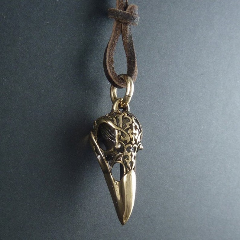 May include: A gold-toned pendant necklace featuring a detailed, ornate bird skull design. The pendant is attached to a brown leather cord.
