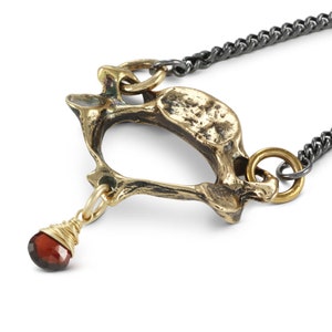 May include: A gold-toned necklace with a pendant shaped like a bone. The pendant is attached to a black chain. A small red gemstone hangs from the bottom of the pendant.