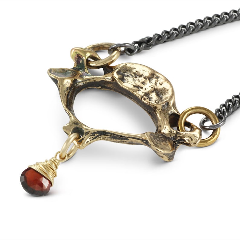 May include: A gold-toned necklace with a pendant shaped like a bone. The pendant is attached to a black chain. A small red gemstone hangs from the bottom of the pendant.