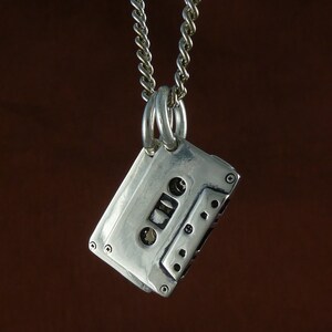 May include: A silver cassette tape pendant hangs from a silver chain. The pendant is a detailed miniature of a classic audio cassette, complete with screw details and a textured surface. The chain is a twisted link style.