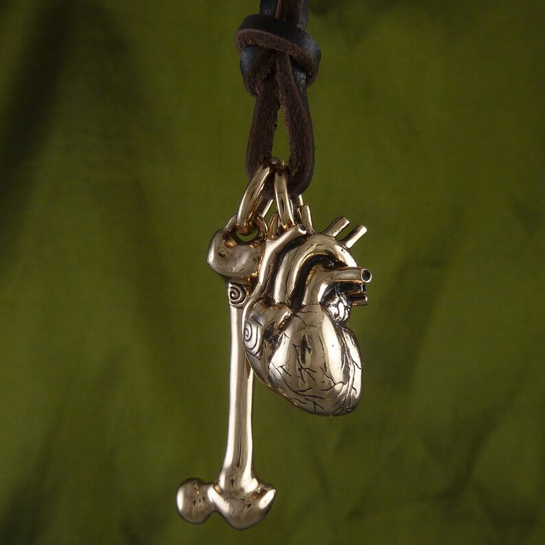 May include: A gold-toned necklace with a detailed anatomical heart and bone pendant. The necklace is made of brown leather cord.