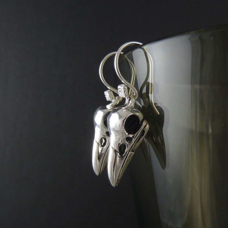 May include: Silver earrings with a detailed skull design. The earrings are hanging from a glass surface.