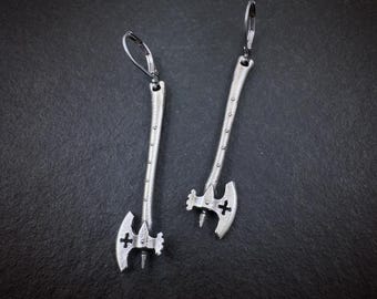 May include: A pair of silver-toned metal drop earrings shaped like battle axes. Each earring features a long, slender handle with a series of small dots and a curved axe head with a cutout cross symbol. The earrings include lever-back closures.