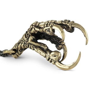 Puede incluir: Un colgante de garra de color bronce con garras afiladas. La garra está detallada con crestas texturizadas y está unida a una fina cadena. El colgante es un accesorio de temática gótica o fantástica.