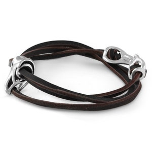 Pode incluir: Um bracelete de couro castanho com detalhes em metal prateado. O bracelete é feito de três tiras de couro e tem um fecho prateado.