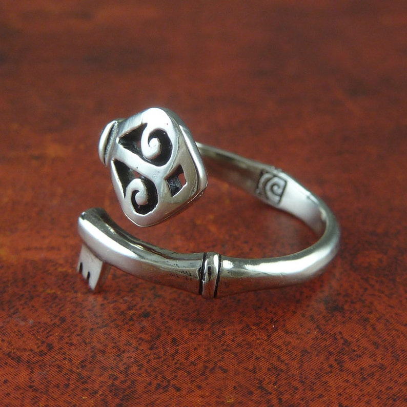 May include: A silver key-shaped ring with a decorative design on the key head. The ring is open and the key head is facing the viewer.