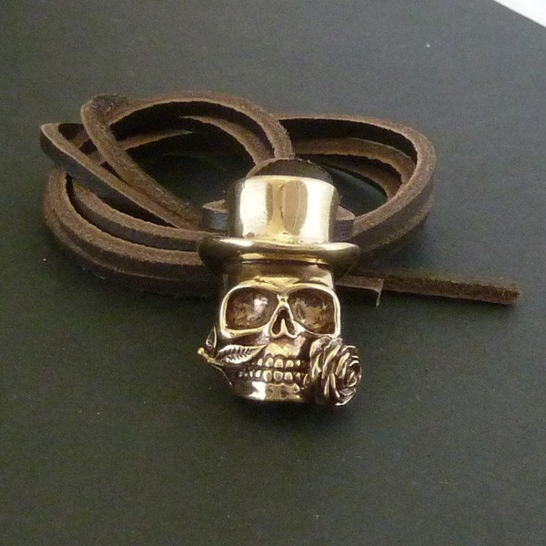 May include: A gold-colored skull pendant with a top hat and a rose in its mouth. The pendant is on a brown leather cord.