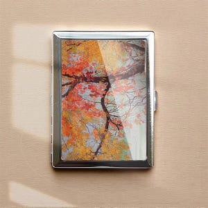 May include: A silver metal cigarette case featuring a vibrant autumn scene. The image showcases a tree with branches and colorful leaves in shades of red, orange, and yellow. The case has a rectangular shape with a hinged closure.