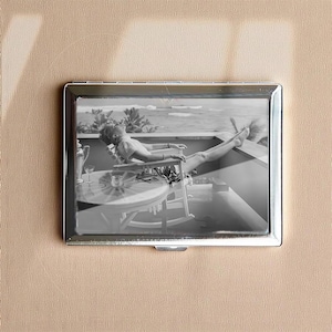 Może przedstawiać: Srebrne metalowe etui na papierosy ze zdjęciem w czerni i bieli osoby relaksującej się na balkonie. Osoba ta leży w fotelu z wyciągniętymi nogami, ciesząc się widokiem na ocean.