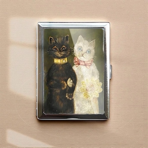May include: A silver metal cigarette case featuring a vintage painting of two cats. One cat is black with a gold collar, and the other is white with a pink bow and a bouquet of flowers.