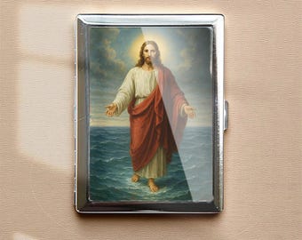 Puede incluir: Un estuche de cigarrillos de metal plateado con una imagen religiosa. La imagen representa una figura con pelo largo y barba, vestida con una túnica blanca y una capa roja, de pie sobre el agua con los brazos extendidos. El fondo muestra un cielo nublado.