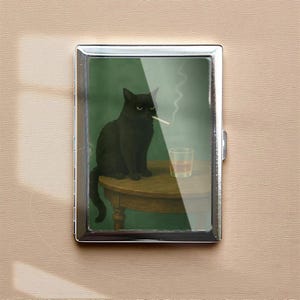 May include: A silver metal cigarette case featuring an illustration of a black cat smoking a cigarette while sitting at a table with a glass of amber liquid. The background is green.