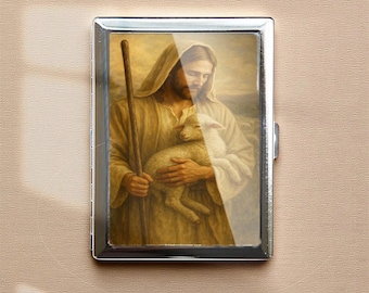 Puede incluir: Un estuche de cigarrillos de metal plateado con una imagen religiosa. La imagen representa una figura con túnicas sosteniendo un cordero, con un bastón en la mano. La obra de arte tiene una paleta de colores cálidos y dorados.
