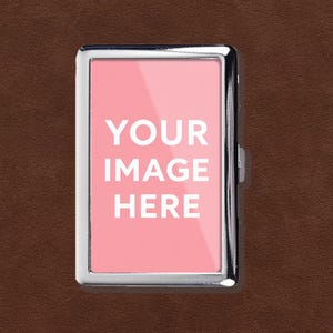 May include: A rectangular, silver-colored cigarette case with a pink insert. The insert features the white text "YOUR IMAGE HERE". The case is set against a brown, textured background.