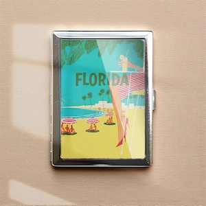 May include: A silver metal cigarette case featuring a vintage Florida beach scene. The artwork depicts a beach with palm trees, people under umbrellas, and the word "FLORIDA" in green. A woman in a pink and white striped skirt and red heels is shown.