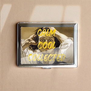 May include: A silver metal cigarette case with a humorous image of a person with their hands on their head. The words "CALM COOL COLLECTED" are written in yellow over the image. The case is set against a neutral background.