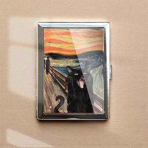 May include: A silver metal cigarette case featuring a parody of Edvard Munch's "The Scream," with a black cat in place of the figure. The cat is depicted with its mouth open, tongue visible, against a colorful, swirling background.