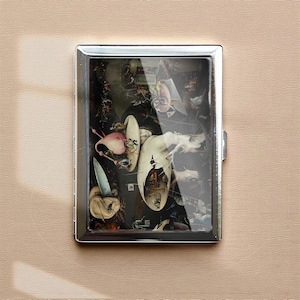 Puede incluir: Un estuche de cigarrillos de metal plateado con una pintura surrealista que presenta figuras y objetos en tonos blancos, rosas y negros. La obra de arte está encerrada en un estuche rectangular con una superficie brillante y reflectante.