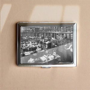 May include: A silver cigarette case featuring a black and white image of a library interior. The image shows people working at desks surrounded by bookshelves filled with books. The case has a rectangular shape.