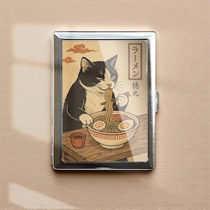May include: A silver metal cigarette case featuring a black and white cat enjoying ramen. The illustration is set against a tan background with Japanese text and a bowl of noodles. A small cup is also visible.