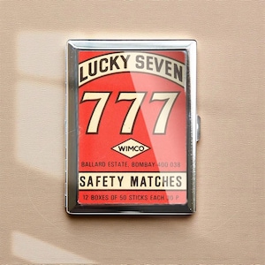 May include: A silver-framed matchbox with a red and white graphic. The graphic reads "LUCKY SEVEN" above the number "777". Below the number is the text "WIMCO" and "SAFETY MATCHES". The matchbox contains 12 boxes of 50 sticks each.