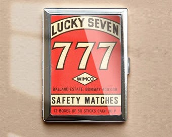 Puede incluir: Una caja de cerillas rectangular con marco plateado y un gráfico rojo y blanco. El gráfico dice "LUCKY SEVEN" encima del número "777". Debajo está el texto "WIMCO" y "SAFETY MATCHES". La caja contiene 12 cajas de 50 cerillas cada una.