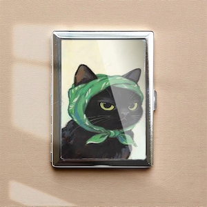May include: A silver metal business card holder featuring a black cat wearing a green headscarf. The cat has yellow eyes and a slightly grumpy expression. The background is a soft yellow.