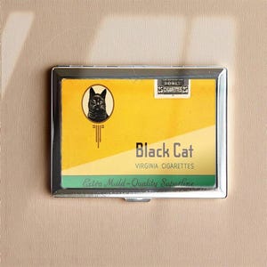 May include: A silver cigarette case with a yellow and green design. The case features the words "Black Cat Virginia Cigarettes" and a graphic of a black cat. The case holds 25 cigarettes.