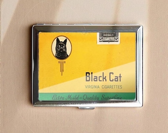 以下が含まれることがあります： 黄色と緑のデザインの銀色のタバコケース。ケースには「Black Cat Virginia Cigarettes」の文字と黒猫のグラフィックが描かれています。ケースには25本のタバコが入っています。