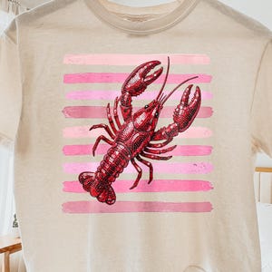 May include: Cream-colored t-shirt featuring a red lobster graphic on a pink striped background. The lobster is detailed with textured shading. The shirt is hanging on a wooden hanger.