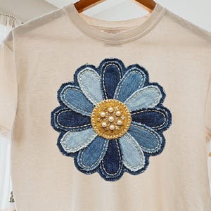 Puede incluir: Una camiseta color crema con una gran flor de mezclilla aplicada. La flor presenta diferentes tonos de pétalos de mezclilla azul y un centro dorado adornado con perlas de imitación. La camiseta está colgada en una percha de madera.