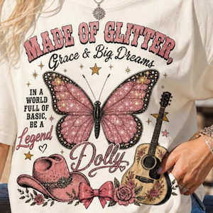 May include: A cream-colored t-shirt featuring a graphic print with the text "MADE OF GLITTER, Grace & Big Dreams, IN A WORLD FULL OF BASIC, BE A Legend Dolly". The design includes a pink glitter butterfly, a cowboy hat, a guitar, and roses.