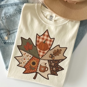 May include: A cream-coloured t-shirt featuring a large maple leaf graphic composed of various autumn-themed patterns including a pumpkin, sunflower, acorn, and a mug of coffee. The shirt is paired with light blue denim jeans and a tan wide-brim hat.