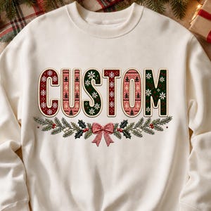 Könnte beinhalten: Ein cremefarbenes Sweatshirt mit Rundhalsausschnitt und dem Wort CUSTOM in großen Blockbuchstaben. Die Buchstaben sind mit roten und grünen Mustern aus Schneeflocken und Tannenbäumen gefüllt. Unter dem Text befindet sich eine Girlande mit einer roten Schleife.