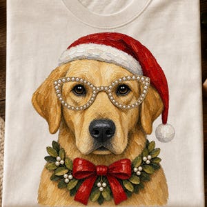 May include: A cream-colored t-shirt featuring a graphic of a golden retriever wearing a red Santa hat, pearl-embellished cat-eye glasses, and a green holly wreath collar with a large red bow. The illustration has a textured, painterly appearance.