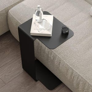 Puede incluir: Una mesa auxiliar negra moderna con un diseño curvo, que sostiene una botella de agua transparente, un libro, un par de gafas y un vaso pequeño. La mesa está colocada junto a un sofá de color claro, creando una estética minimalista.