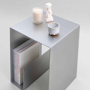 Puede incluir: Una mesa auxiliar moderna de metal gris con un revistero. La superficie de la mesa tiene una vela, una pequeña escultura y una taza de cerámica. Las revistas en el estante incluyen "Vogue Beauty". La mesa tiene un diseño minimalista.